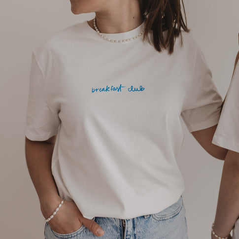 Frau trägt weißes T-Shirt mit „breakfast club“-Print in Blau – moderner Look mit Perlenkette und Jeans.