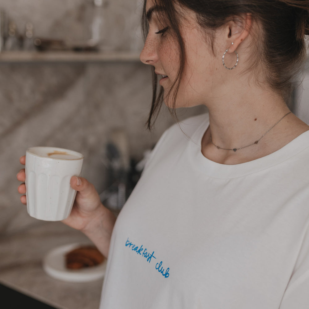 Frau mit weißem Shirt und blauem „breakfast club“-Aufdruck trinkt Kaffee.