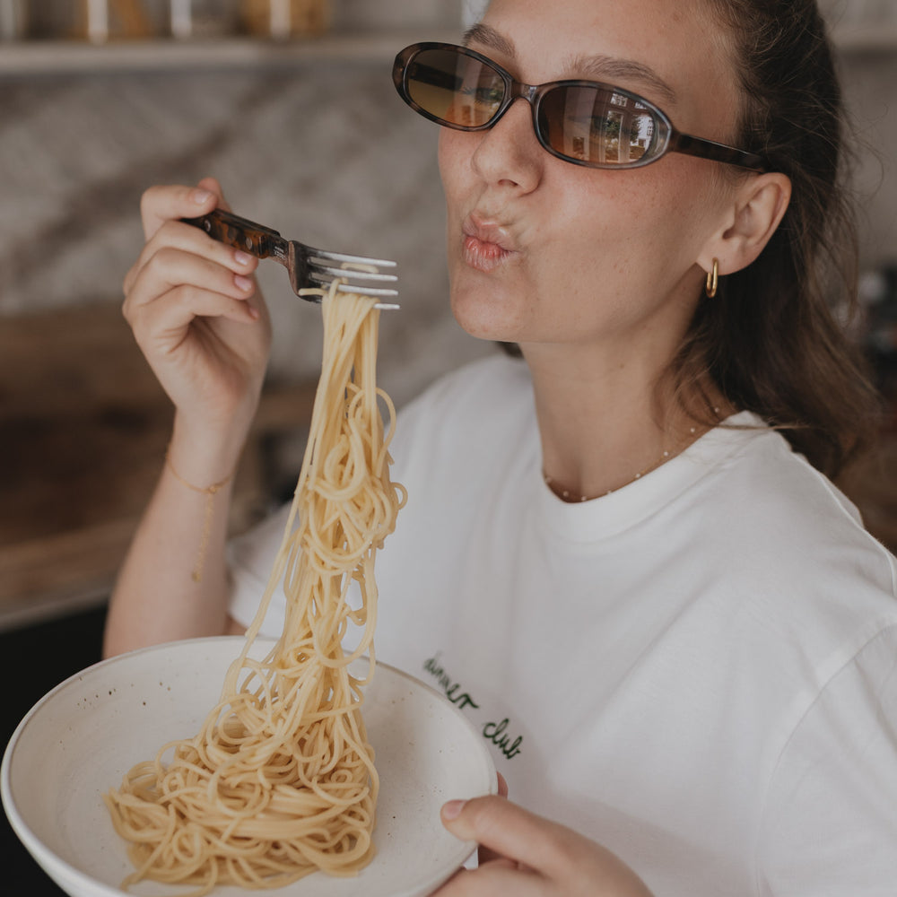 Frau mit Spaghetti und weißem Dinner Club Shirt, humorvolle Essensszene in der Küche.