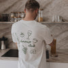 Rückansicht eines Mannes mit weißem Dinner Club Shirt und grünen Food-Icons in moderner Küche.