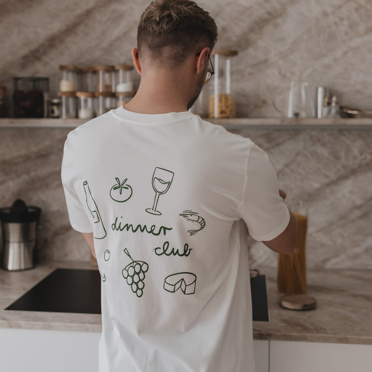 Rückansicht eines Mannes mit weißem Dinner Club Shirt und grünen Food-Icons in moderner Küche.