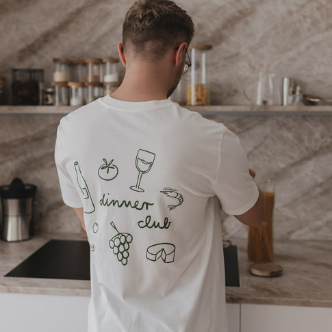 Rückansicht eines Mannes mit weißem Dinner Club Shirt und grünen Food-Icons in moderner Küche.