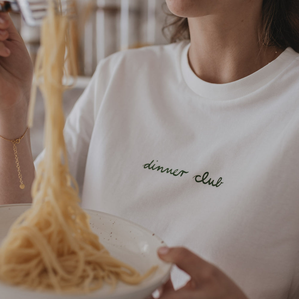 Weißes T-Shirt mit grünem „dinner club“-Schriftzug, getragen beim Servieren von Spaghetti.