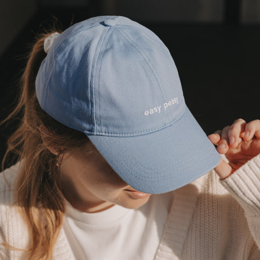 Hellblaue Cap mit weißer Stickerei „easy peasy“, getragen von Frau im Sonnenlicht