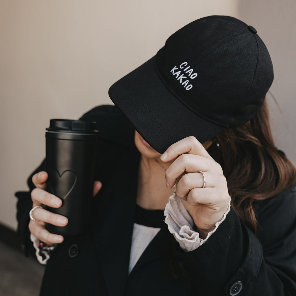 Frau mit schwarzer Cap „Ciao Kakao“ und schwarzem Coffee-to-go Becher