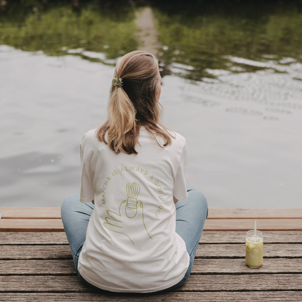 Frau sitzt am Wasser mit weißem Matcha T-Shirt, entspannte Outdoor-Szene