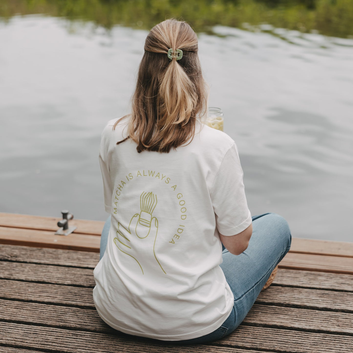 Frau mit weißem Matcha T-Shirt am See, Rückenansicht mit Getränk