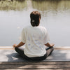 Frau meditiert am See auf Yogamatte, trägt weißes T-Shirt „go with the flow“ mit Rückendruck