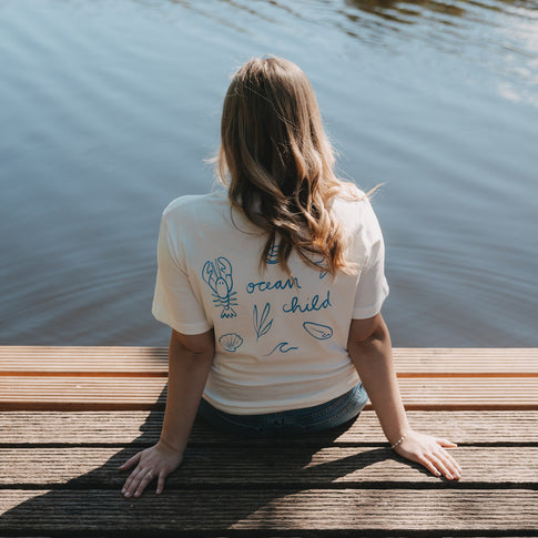 Frau am Wasser mit weißem T-Shirt „Ocean Child“ und blauem Rückenprint, entspannt am Steg