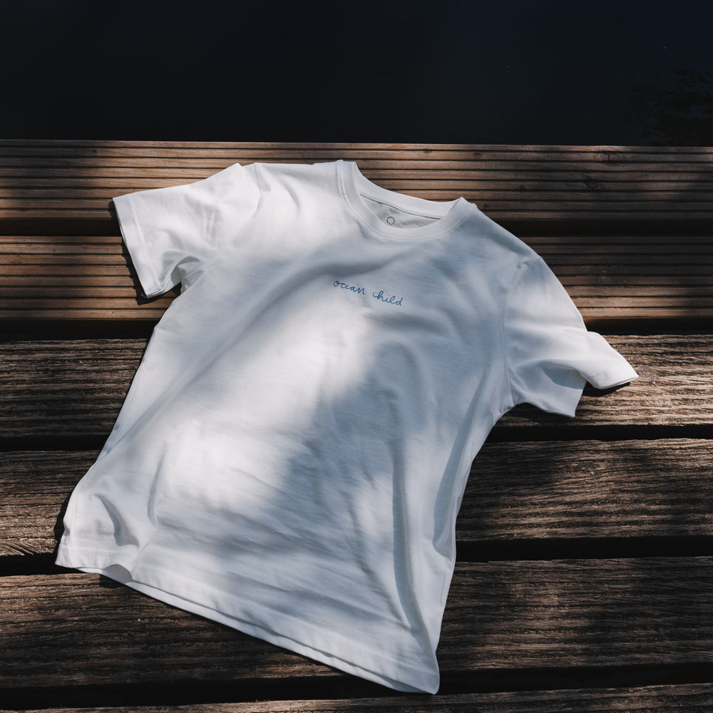 Weißes „Ocean Child“ T-Shirt flach auf Holzsteg liegend, minimalistische Produktansicht