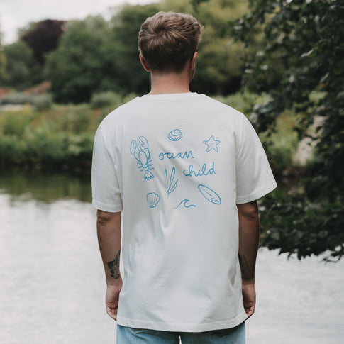 Rückenansicht Mann im weißen „Ocean Child“ T-Shirt mit blauem Print am Wasser