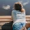 Rückenansicht weißes „Ocean Child“ T-Shirt mit blauem Print, entspannt am Steg liegend