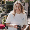 Frau mit Brille hält Kaffeebecher, trägt weißes T-Shirt mit kleinem Coffee-Motiv vorne im Garten