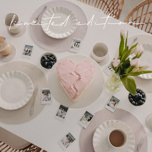 Gedeckter Tisch mit wendbaren Platzsets „Lieblingsmensch“ in Flieder, Herzkuchen, Keramikgeschirr und Blumen.