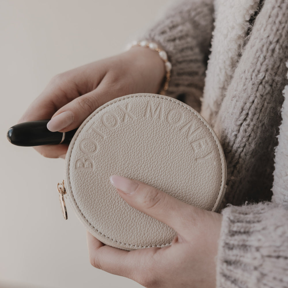 Rundes Portemonnaie mit BOTOX MONEY Prägung, in Händen gehalten, beige Struktur und goldener Reißverschluss.