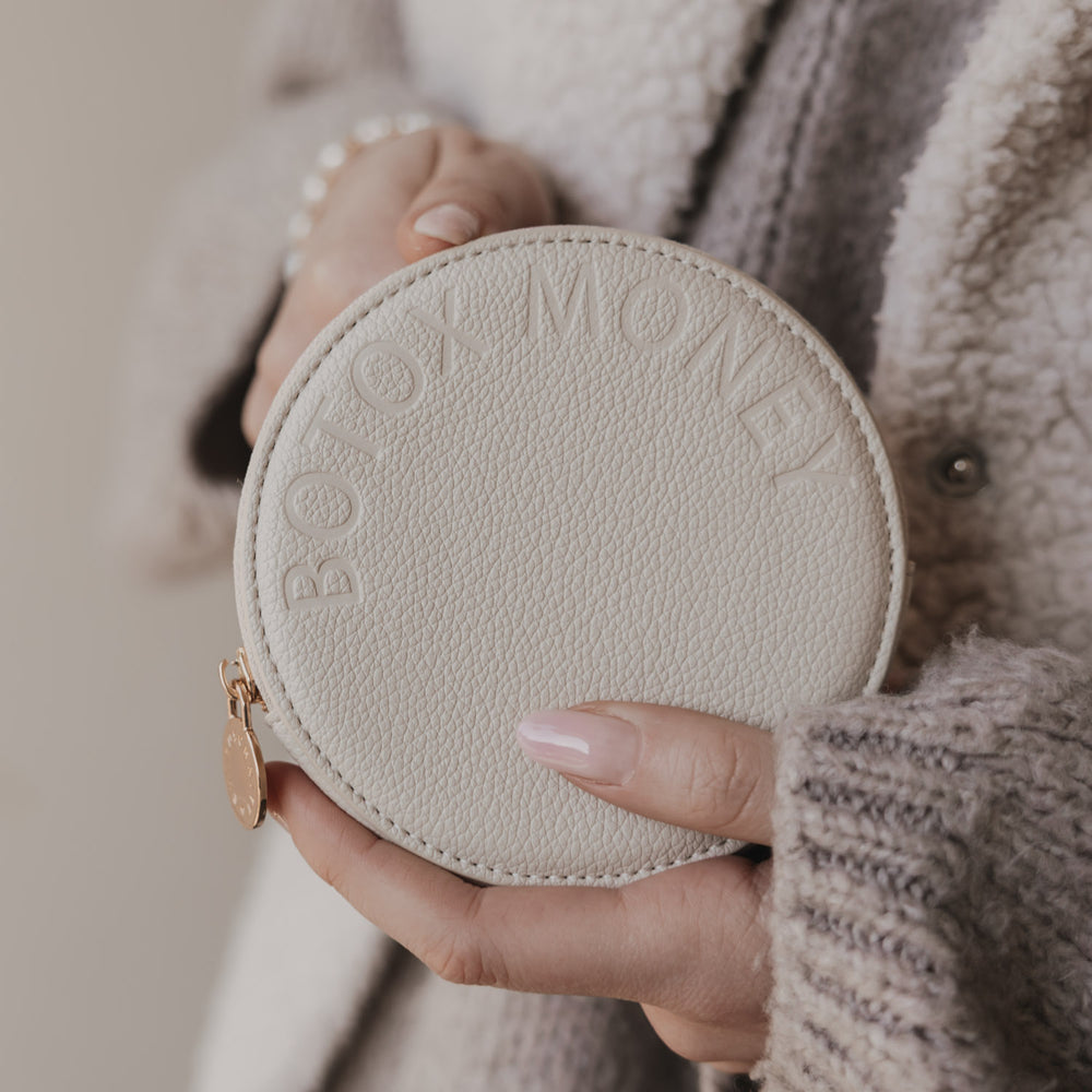 Beiges rundes Portemonnaie mit BOTOX MONEY Prägung, in Händen gehalten, goldener Zipperanhänger.