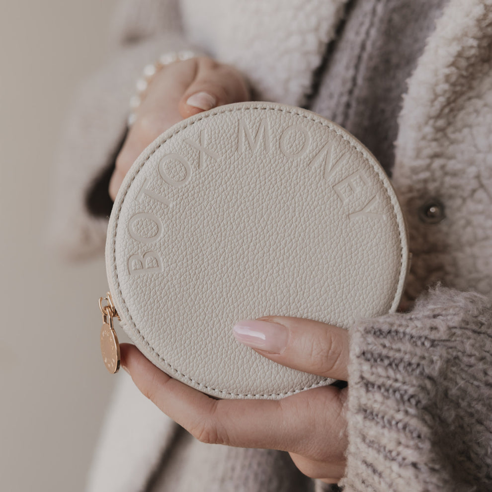 Beiges rundes Portemonnaie mit BOTOX MONEY Prägung, in Händen gehalten, goldener Zipperanhänger.