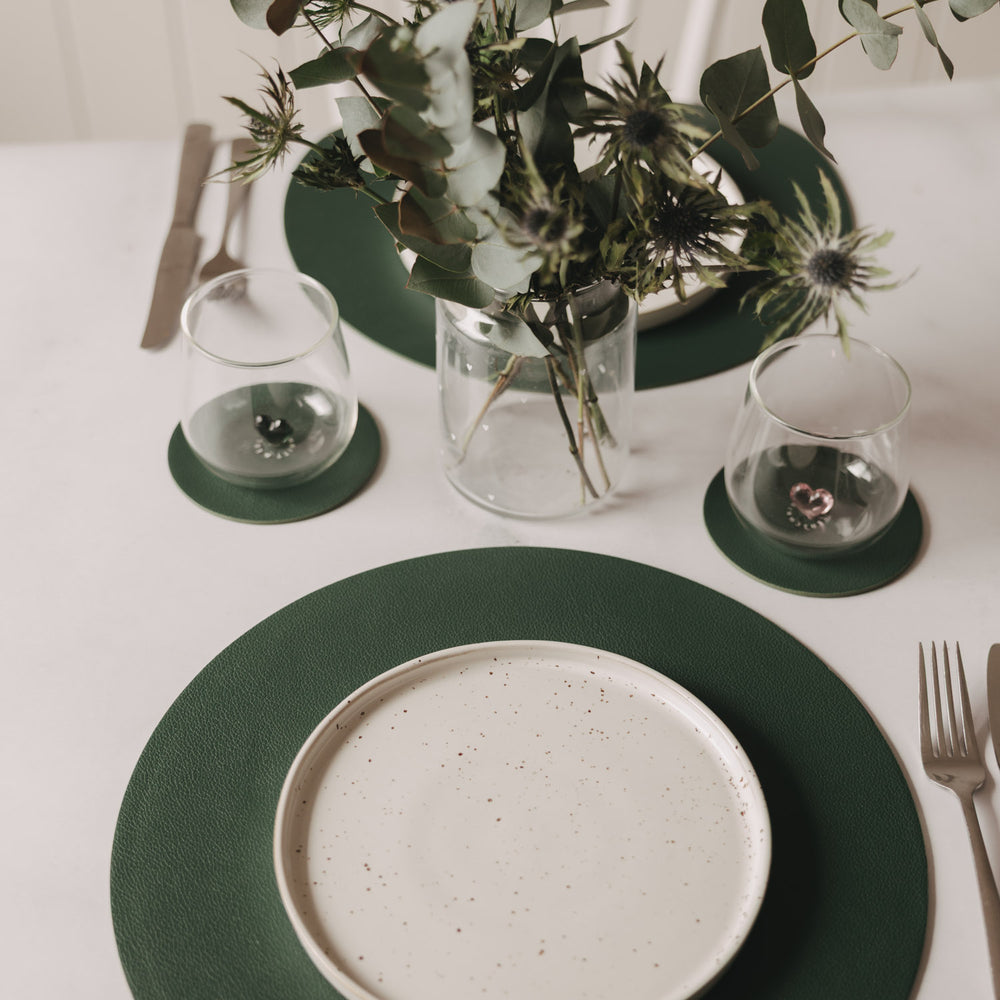 Gedeckter Tisch mit dunkelgrünen Untersetzern, rundem Platzset, Teller und Blumen