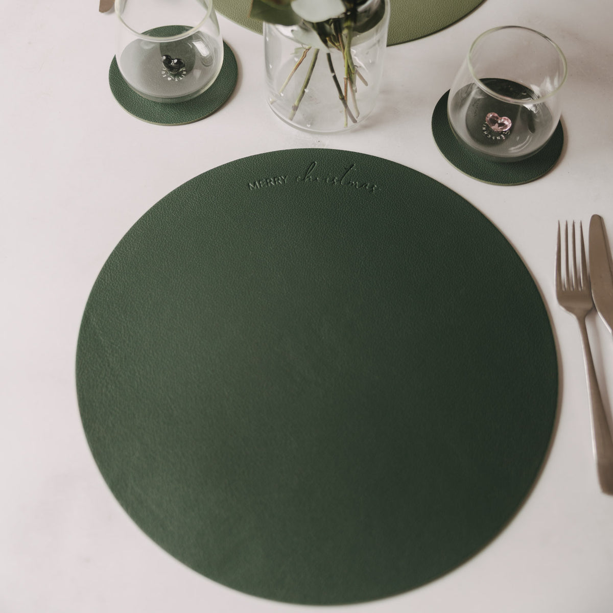 Dunkelgrünes rundes Platzset mit geprägtem Merry-Christmas-Schriftzug auf gedecktem Tisch