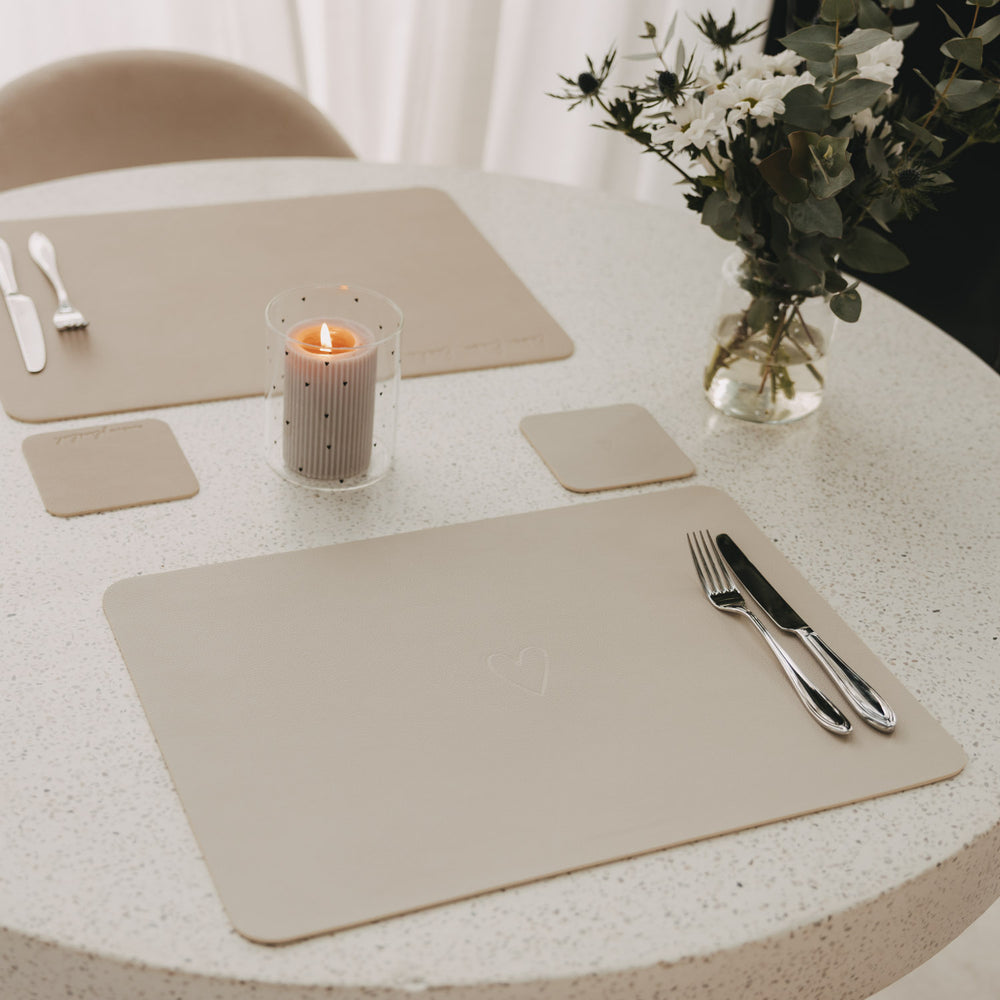 Gedeckter Tisch mit cremefarbenen Platzsets und quadratischen Untersetzern, Kerze und Blumenstrauß