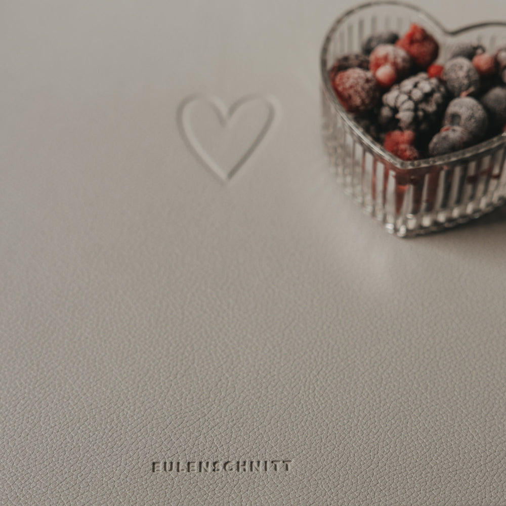 Hellgraues Platzset mit Herzprägung und Eulenschnitt Logo neben Schale mit Beeren