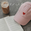 Rosa Cap mit rotem Herz neben Iced Coffee und aufgeschlagenem Buch