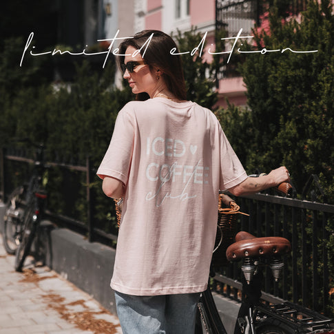 Rückansicht einer Frau im rosafarbenen Oversize-T-Shirt mit weißem „Iced Coffee Club“-Print neben einem Fahrrad.