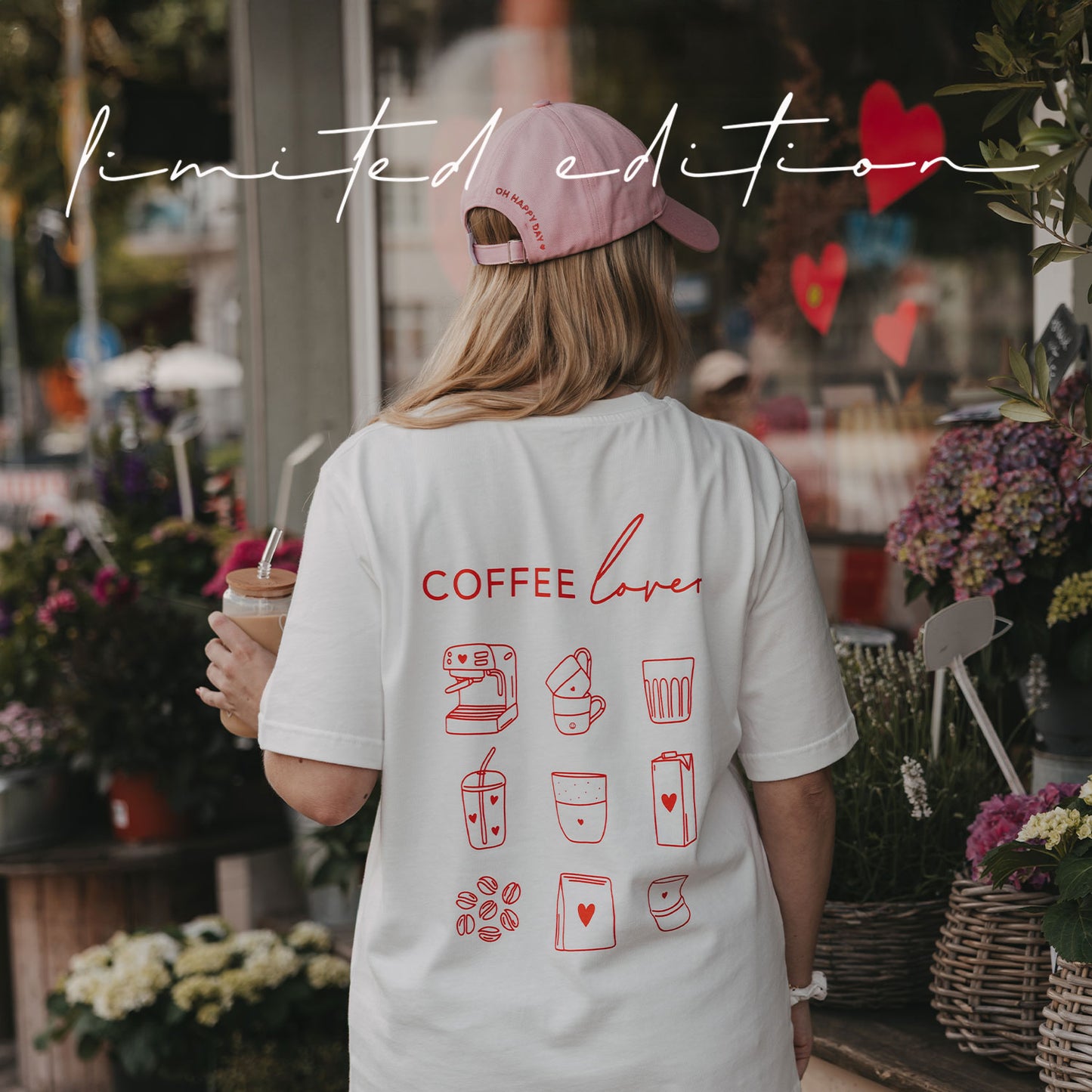 Rückansicht einer Frau im weißen „Coffee Lover“-T-Shirt mit rotem Kaffee-Print und rosa Cap vor einem Blumenladen.