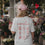 Rückansicht einer Frau im weißen „Coffee Lover“-T-Shirt mit rotem Kaffee-Print und rosa Cap vor einem Blumenladen.