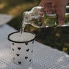 Weißer Emaille-Becher mit schwarzem Herzmuster wird im sommerlichen Picknick-Setting mit Wasser befüllt.