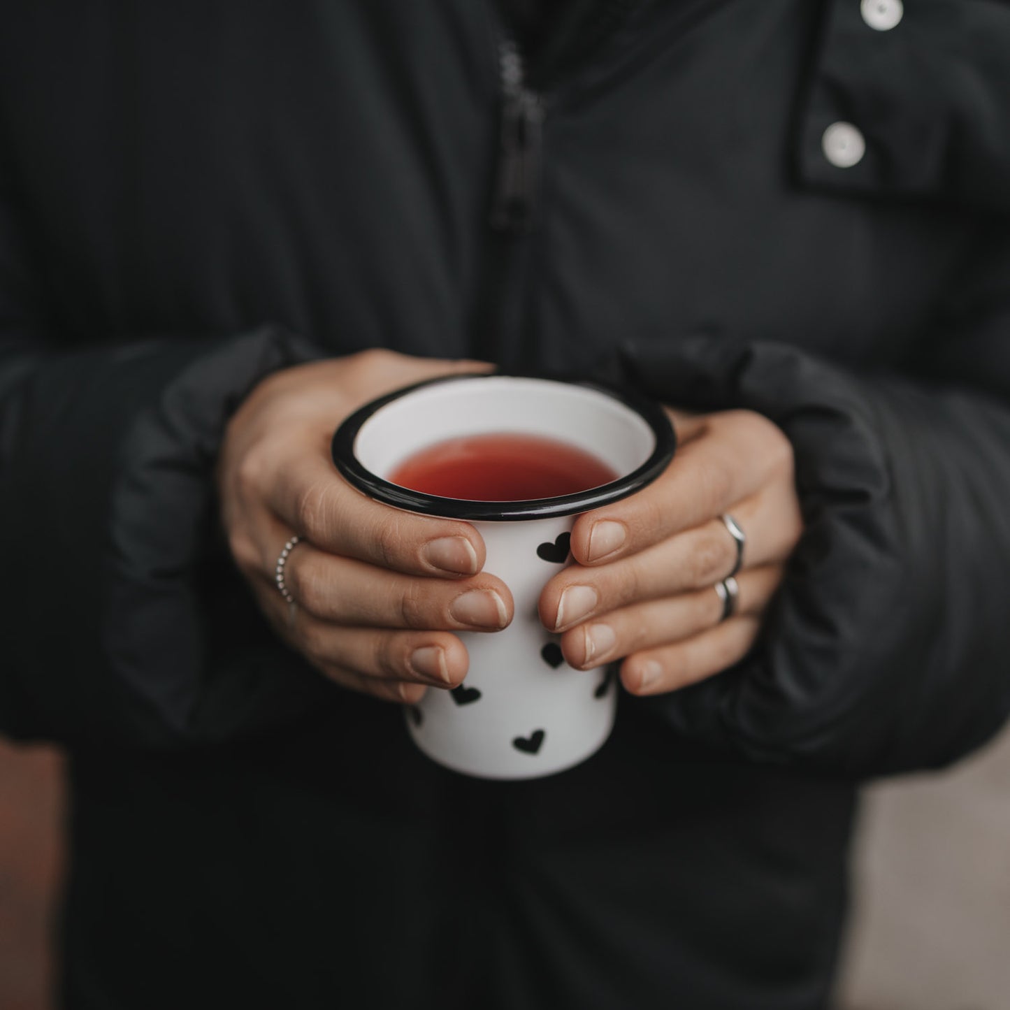 Person hält Emaillebecher mit schwarzen Herzen und warmem Getränk in beiden Händen vor dunkler Jacke.