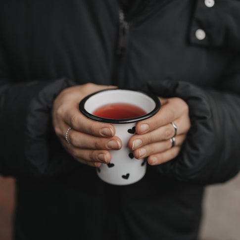 Person hält Emaillebecher mit schwarzen Herzen und warmem Getränk in beiden Händen vor dunkler Jacke.