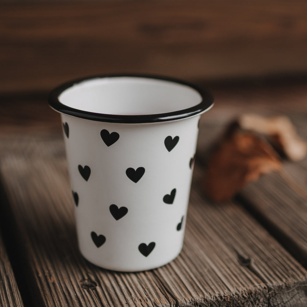 Weißer Emaillebecher mit schwarzen Herzen steht auf Holzoberfläche, herbstliche Deko im Hintergrund.