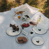 Weißer Emaille-Becher mit großem schwarzem Herz im sommerlichen Picknick-Setting mit passendem Emaille-Geschirr.