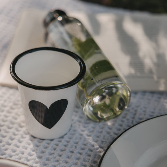 Weißer Emaille-Becher mit großem schwarzem Herz neben Trinkflasche auf einer Picknickdecke.