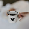 Weiße Emaille-Espressotasse mit schwarzem Herz, gefüllt mit Espresso, wird am Henkel gehalten.