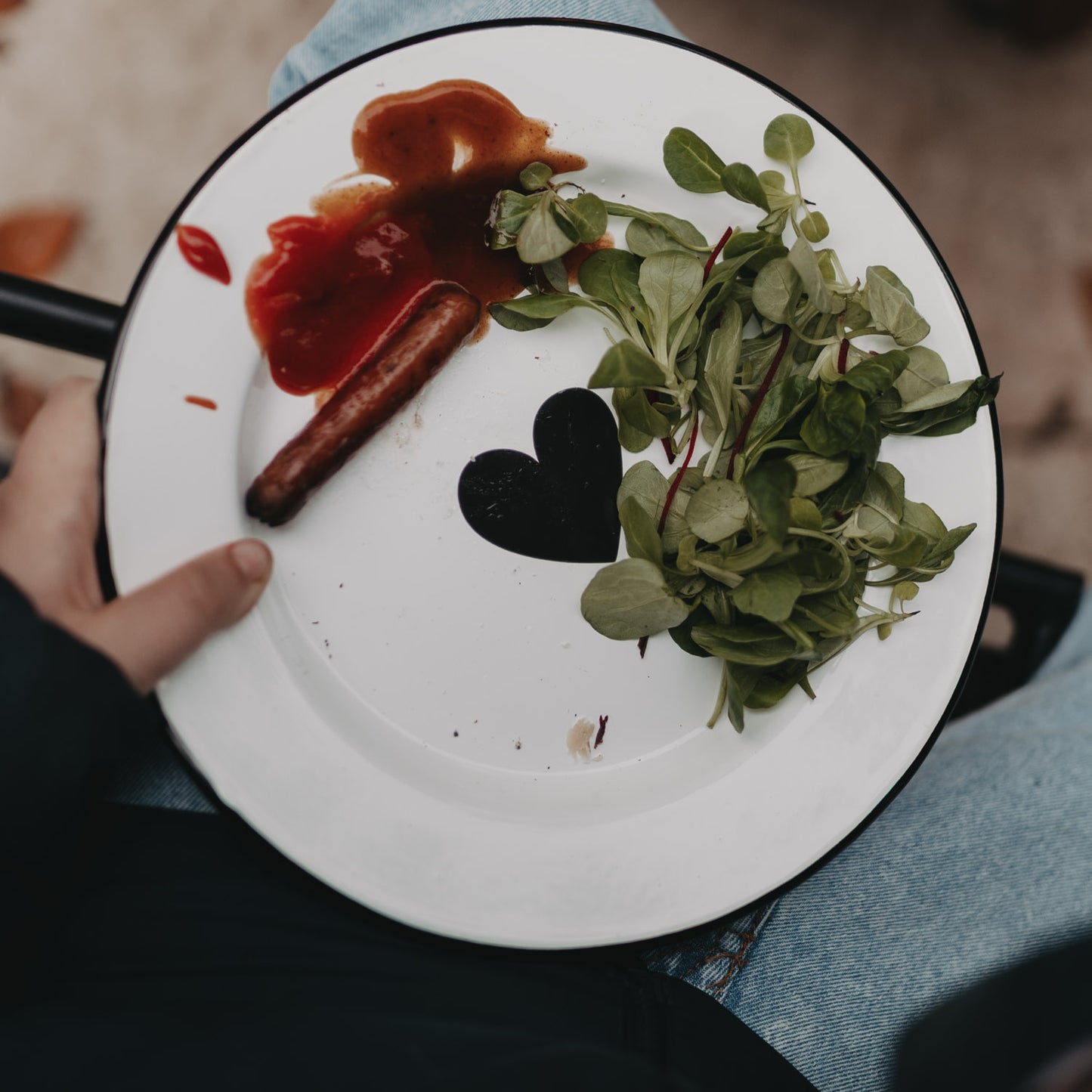 Emaille Teller Herz mit Salat, Soße und Würstchen auf dem Schoß – ideal für Picknick oder Grillabend.