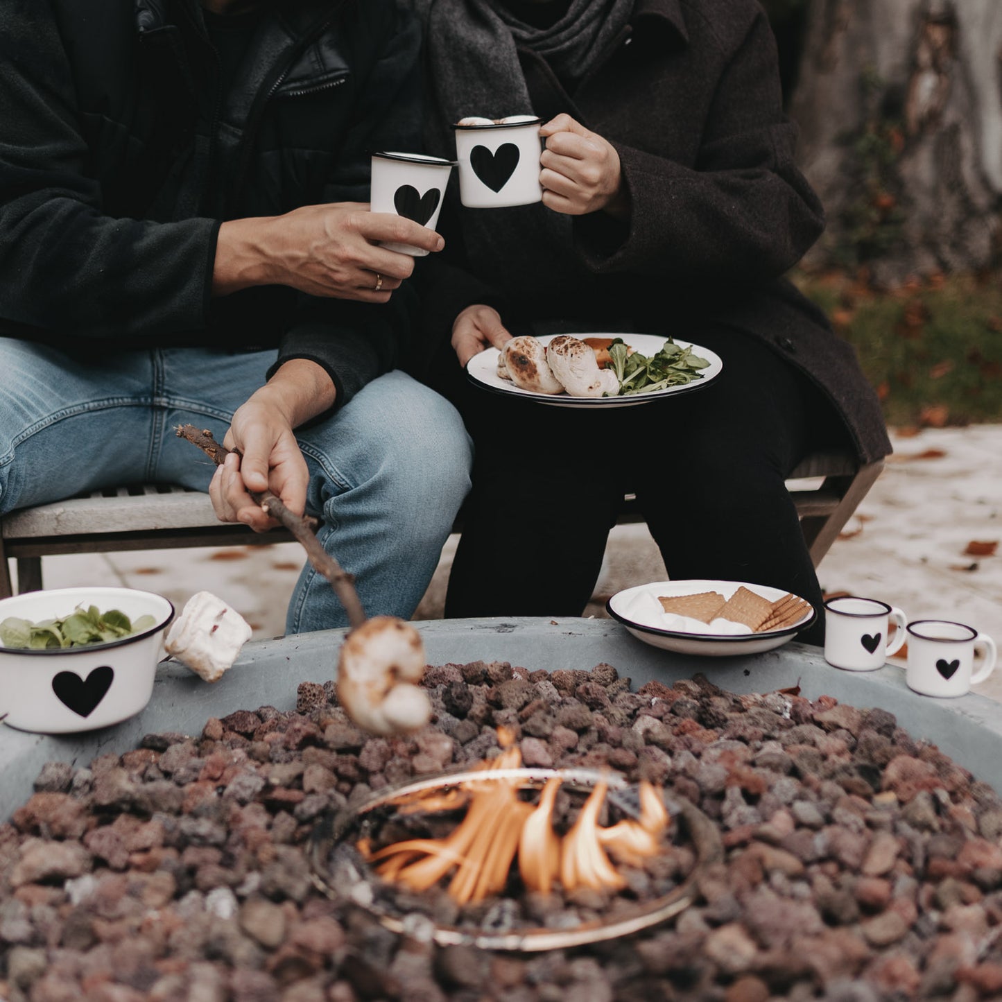 Zwei Personen genießen Essen und Getränke mit Emaille Geschirr Herz am Lagerfeuer – stimmungsvolles Outdoor-Set.