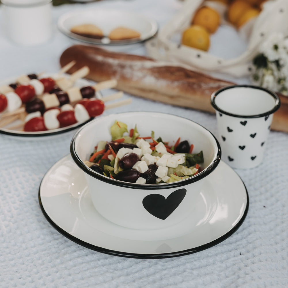 Weiße Emaille-Schale mit schwarzem Herz auf passendem Emaille-Teller, gefüllt mit Salat, im sommerlichen Picknick-Setting.