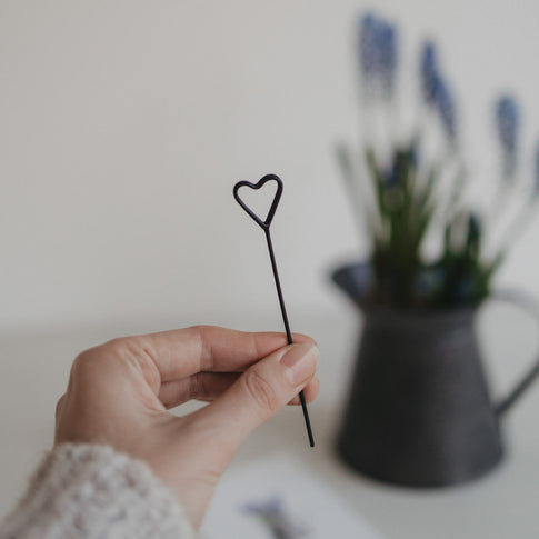 Schwarzer Herz-Stecker wird in der Hand gehalten, unscharfe Blumen im Hintergrund.