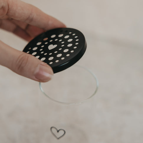 Hand hebt schwarzen Lochdeckel vom Mini-Blumenhalter aus Glas mit Herzmotiv, moderne Deko von Eulenschnitt.