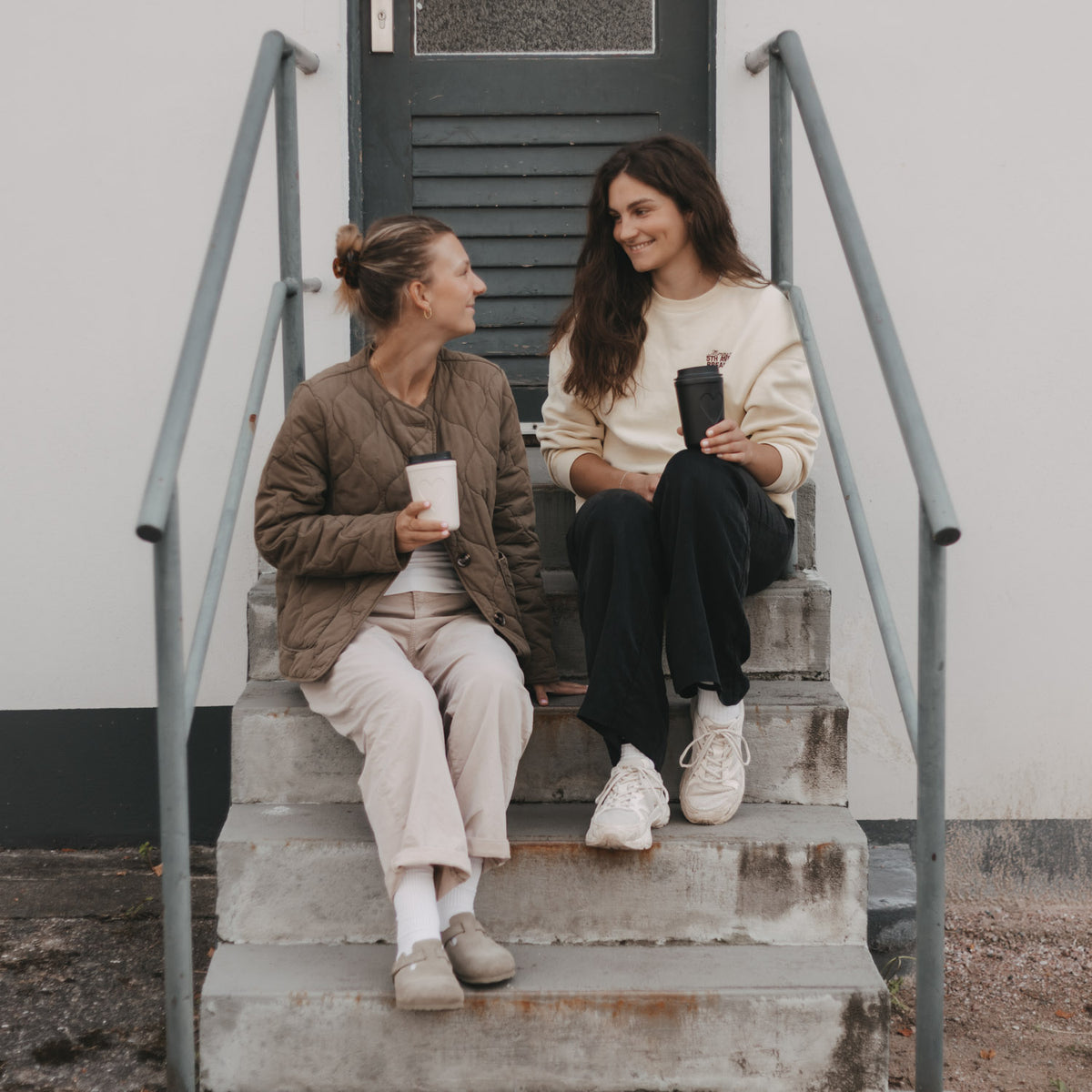 Zwei Frauen sitzen mit schwarzen und weißen To-Go-Bechern mit Herzmotiv von Eulenschnitt auf einer Treppe