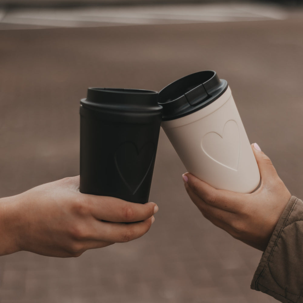 Schwarzer und cremefarbener To-Go-Becher mit Herzmotiv werden zum Anstoßen zusammengehalten