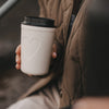 Cremefarbener To-Go-Becher mit Herzmotiv und schwarzem Deckel in der Hand einer Frau