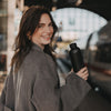 Lächelnde Frau hält eine schwarze Trinkflasche mit Herzmotiv und Schraubverschluss unterwegs am Bahnhof.