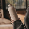 Frau im grauen Mantel hält eine cremefarbene 1000 ml Trinkflasche mit geprägtem Herzmotiv in der Hand.