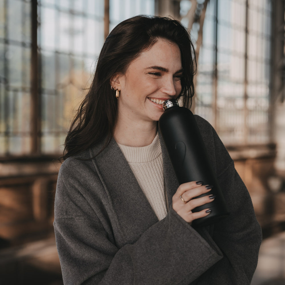 Lächelnde Frau im grauen Mantel hält eine geöffnete schwarze Trinkflasche mit Herzmotiv nah an ihr Gesicht.