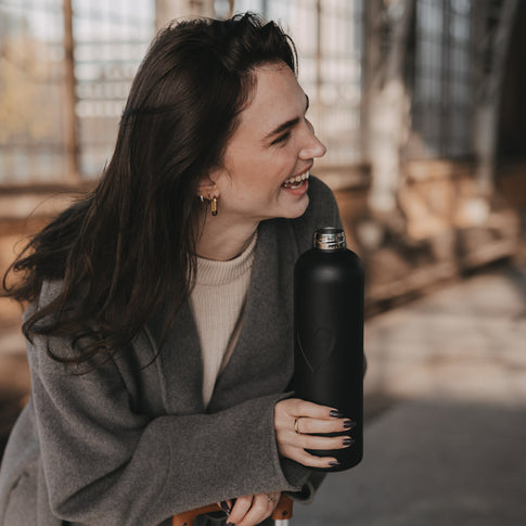 Lachende Frau hält eine schwarze 1000 ml Trinkflasche mit geprägtem Herzmotiv und offenem Flaschenhals.