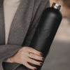 Detailaufnahme einer mattschwarzen Trinkflasche mit geprägtem Herzmotiv und silbernem Flaschenhals in einer Hand.