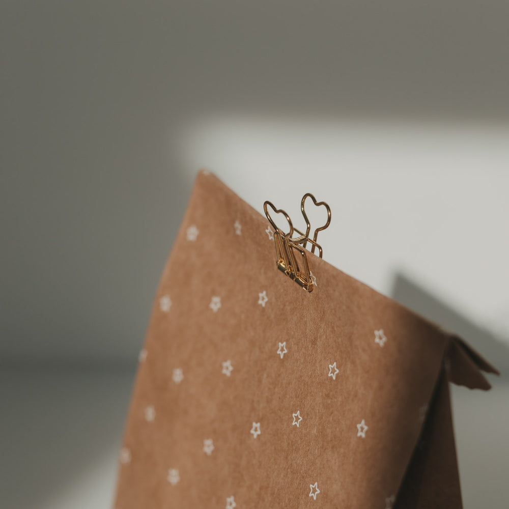 Nahaufnahme eines goldenen Foldback-Clips mit Herzmotiv an einer Geschenktüte aus Kraftpapier mit Sternenmuster.